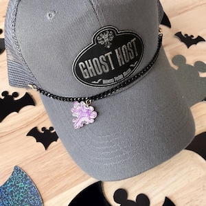 May include: A grey trucker hat featuring a black oval patch with the text GHOST HOST and Welcome Foolish Mortals. A thin black metal chain is attached to the sides of the hat, featuring a small, translucent, light purple, glittery ghost charm.
