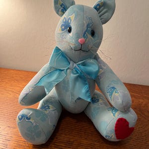 May include: A light blue plush cat with butterfly and floral patterns. The cat has a pink nose, whiskers, and a blue satin bow. A red heart is sewn on one paw. The cat is sitting on a wooden surface.