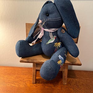 May include: A denim blue bunny rabbit plush toy with embroidered flowers and a hummingbird. The bunny is sitting on a small wooden stool.