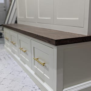 May include: White painted wooden cabinet with a dark wood top and gold drawer pulls. The cabinet has a bench seat and multiple drawers.