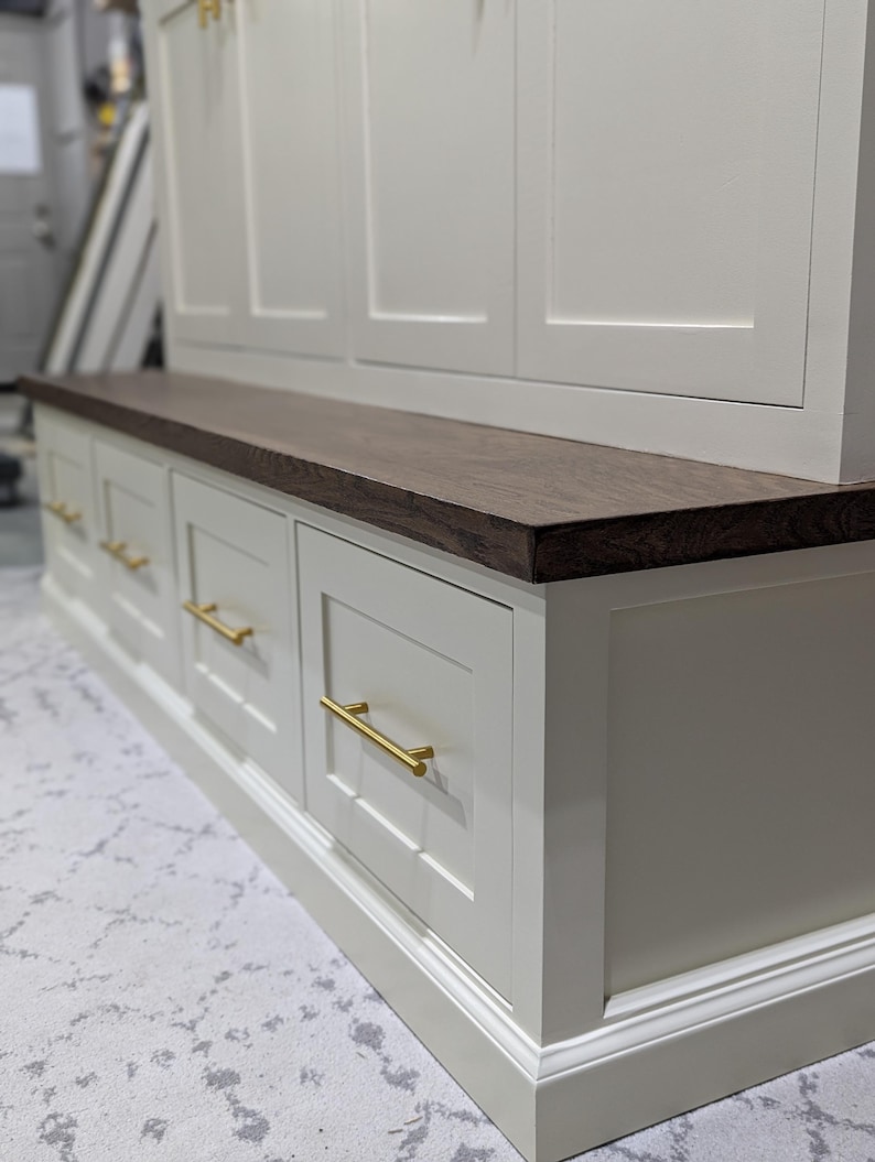 May include: White painted wooden cabinet with a dark wood top and gold drawer pulls. The cabinet has a bench seat and multiple drawers.