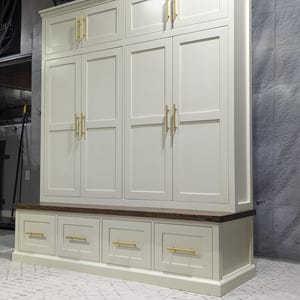 May include: A white painted wooden storage cabinet with a bench and drawers. The cabinet has six doors with gold-toned hardware. The bench has four drawers with gold-toned hardware.