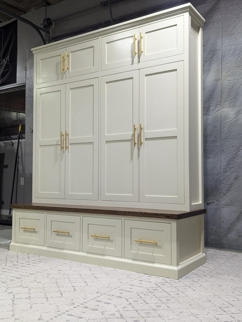 May include: A white painted wooden storage cabinet with a bench and drawers. The cabinet has six doors with gold-toned hardware. The bench has four drawers with gold-toned hardware.