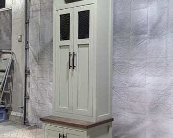 May include: A tall, light green wooden cabinet with two doors and a shelf. The doors have black metal mesh inserts and black metal handles. The cabinet has a lower storage area with two doors and black metal handles. The cabinet is sitting on a wooden base.