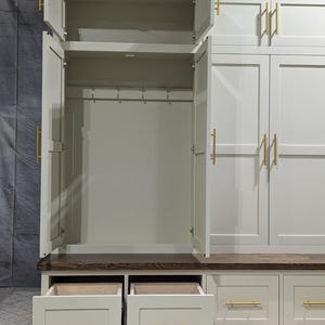 May include: White wooden hallway cabinet with two doors and a bench. The cabinet has a shelf and hooks inside. The bench has two drawers. The cabinet has gold hardware.