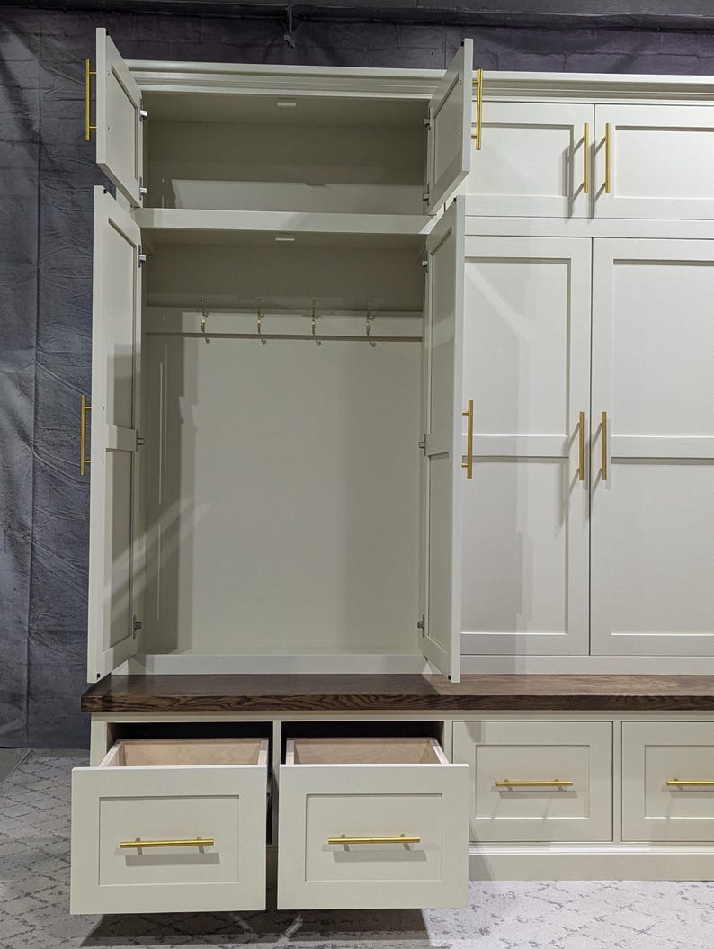 May include: White wooden hallway cabinet with two doors and a bench. The cabinet has a shelf and hooks inside. The bench has two drawers. The cabinet has gold hardware.