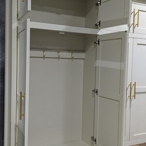 May include: White wooden cabinet with two doors and a shelf. The doors are open, revealing a shelf and hooks inside. The cabinet has gold-coloured hardware.
