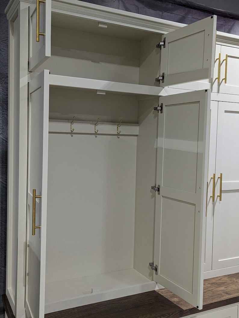 May include: White wooden cabinet with two doors and a shelf. The doors are open, revealing a shelf and hooks inside. The cabinet has gold-coloured hardware.