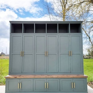 May include: A gray wooden storage cabinet with a bench seat. The cabinet has six doors with gold handles and a top shelf with four compartments. The bench seat has three drawers with gold handles.