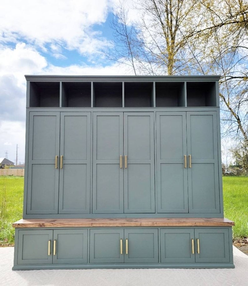 May include: A gray wooden storage cabinet with a bench seat. The cabinet has six doors with gold handles and a top shelf with four compartments. The bench seat has three drawers with gold handles.