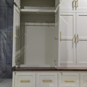 May include: A white painted wooden storage cabinet with a bench seat and drawers. The cabinet has two doors with gold handles and a shelf inside. The bench seat has a dark wood top and three drawers with gold handles.