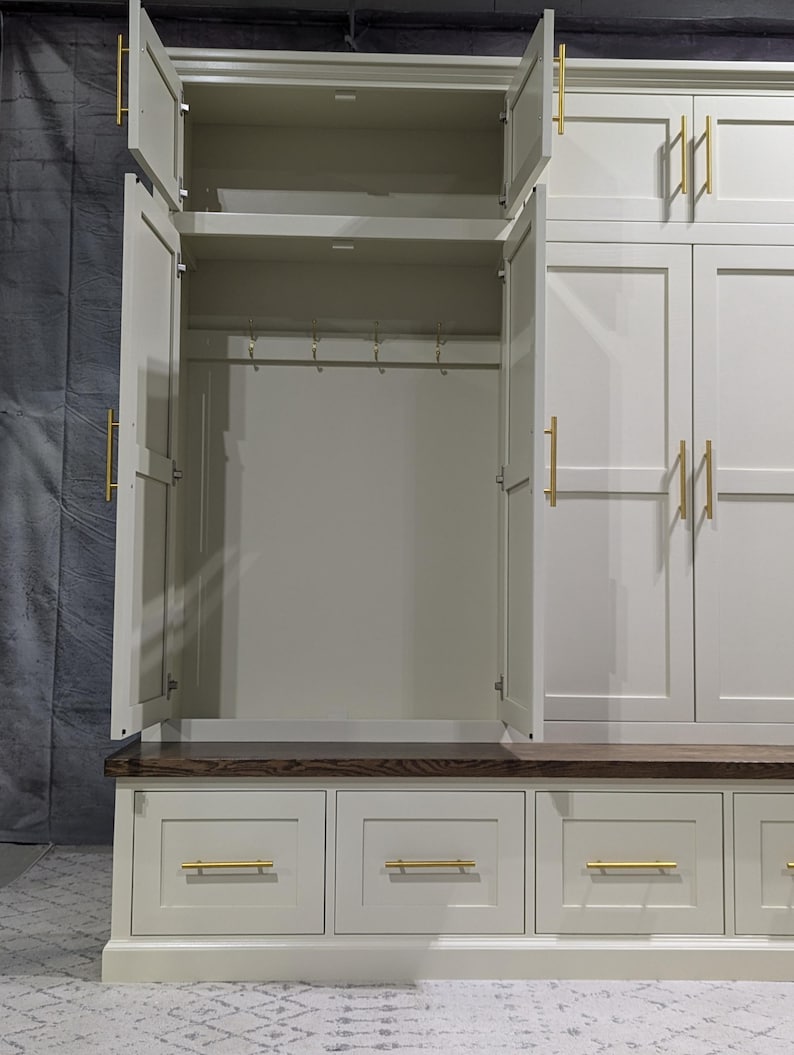 May include: A white painted wooden storage cabinet with a bench seat and drawers. The cabinet has two doors with gold handles and a shelf inside. The bench seat has a dark wood top and three drawers with gold handles.