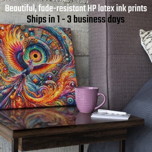 May include: A colorful canvas print featuring a stylized phoenix with a large eye in the center. The print is framed and displayed on a wooden table with a purple mug and a notebook.
