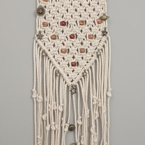 May include: Cream-coloured macrame wall hanging with wooden dowel and hanging cord. The woven design features small wooden beads and decorative pins with celestial and geometric shapes. Knotted fringe hangs below.