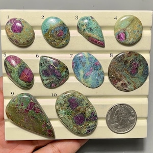 Könnte beinhalten: Eine Sammlung von zehn polierten Rubin-Zoisit-Edelsteinen in verschiedenen Formen, darunter oval, tropfenförmig und rund. Die Steine zeigen eine Mischung aus Grün-, Blaugrün- und Pink-Rot-Tönen. Eine US-Viertel-Dollar-Münze dient als Größenreferenz.