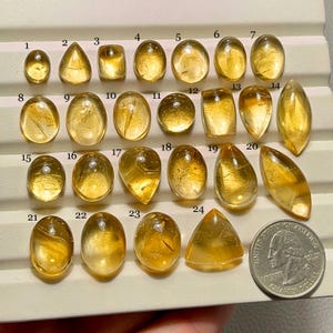 May include: A collection of twenty-four polished, golden-yellow gemstones of various shapes, including oval, teardrop, and rectangular. A US quarter coin is included for size comparison. The gemstones are numbered 1 through 24.