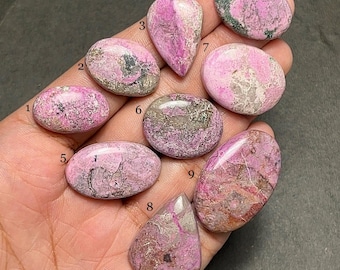 May include: A collection of nine polished rhodonite gemstones in various shapes, including oval, teardrop, and rounded forms. The stones display a vibrant pink hue with dark gray and black veining, showcasing natural patterns. The gemstones are held in a hand.