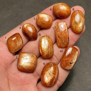 May include: A collection of ten polished sunstone gemstones in various shapes, including oval, rectangular, and square. The stones display a warm orange hue with shimmering inclusions, reflecting light. The gemstones are held in a hand.