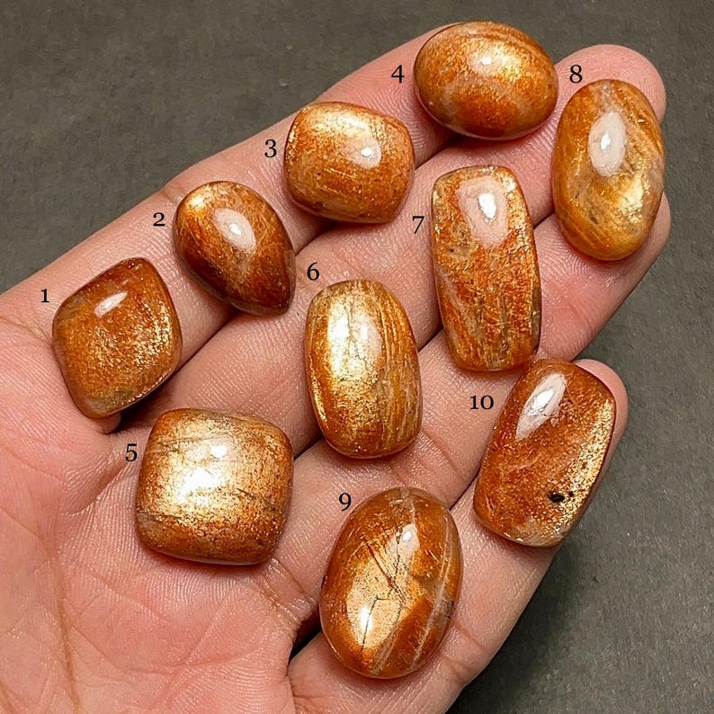 May include: A collection of ten polished sunstone gemstones in various shapes, including oval, rectangular, and square. The stones display a warm orange hue with shimmering inclusions, reflecting light. The gemstones are held in a hand.