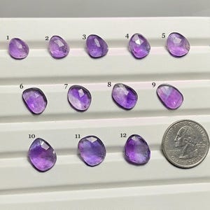 May include: Twelve faceted amethyst gemstones in various shapes and sizes are displayed on a white surface. The gemstones range in color from light to deep purple. A US quarter coin is included for size reference.
