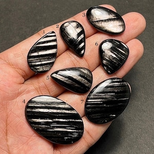 Puede incluir: Se muestran siete piedras preciosas de obsidiana negra pulida con inclusiones plateadas. Las piedras tienen varias formas, incluyendo lágrimas, óvalos y formas irregulares. Las piedras preciosas se sostienen en una mano sobre un fondo oscuro.