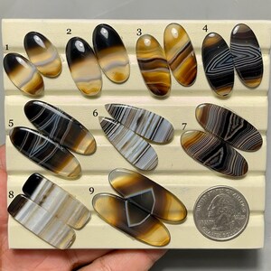 May include: A collection of nine pairs of polished agate gemstones in various colors and patterns. The stones are oval-shaped and displayed on a cream-colored surface. A US quarter coin is included for size reference. The gemstones feature brown, white, black, and yellow hues.