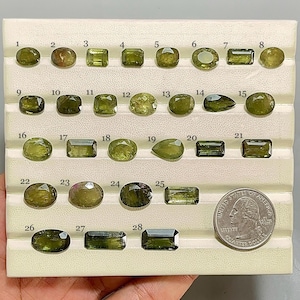 Puede incluir: Una colección de 28 gemas verdes de diversas formas, incluyendo óvalos, rectángulos y piedras en forma de pera. Se incluye una moneda de cuarto de dólar para comparar el tamaño. Las gemas están dispuestas en una bandeja blanca.