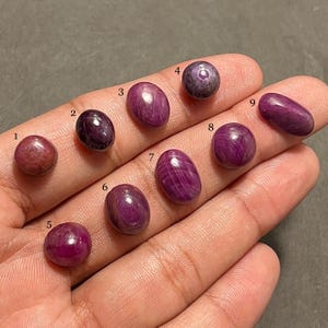 Könnte beinhalten: Eine Sammlung von neun polierten Edelsteinen in verschiedenen Formen und Farbtönen von Rot und Lila. Die Edelsteine sind oval, rund und länglich, mit einer glatten, reflektierenden Oberfläche. Die Edelsteine sind von 1 bis 9 nummeriert.