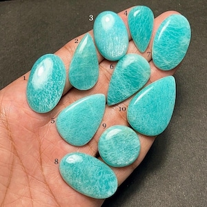 Op de afbeelding: Een verzameling van tien Amazoniet edelstenen in verschillende vormen, waaronder ovaal en druppelvormig. De stenen vertonen een levendige turquoise kleur met subtiele witte adering, die natuurlijke patronen laten zien. De gepolijste edelstenen zijn glad en reflecteren licht. Afmetingen in centimeters.
