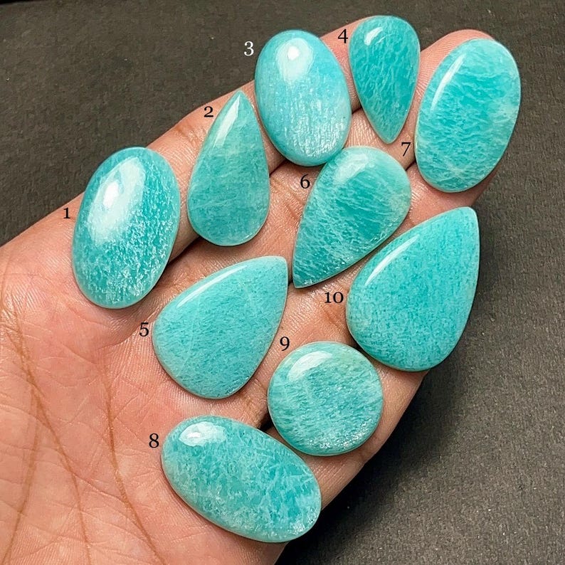 Op de afbeelding: Een verzameling van tien Amazoniet edelstenen in verschillende vormen, waaronder ovaal en druppelvormig. De stenen vertonen een levendige turquoise kleur met subtiele witte adering, die natuurlijke patronen laten zien. De gepolijste edelstenen zijn glad en reflecteren licht. Afmetingen in centimeters.