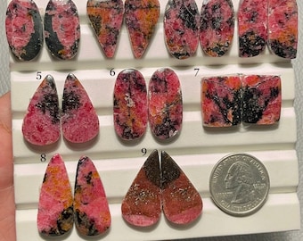 May include: A collection of nine pairs of polished rhodonite gemstones in various shapes, including oval, teardrop, and rectangular. The stones display a mix of pink, black, and orange hues, with a quarter coin for size comparison. The gemstones are set on a white surface.