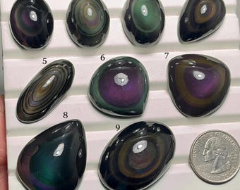 May include: A collection of nine polished obsidian gemstones, each with unique swirling patterns of iridescent colors including purple, green, and gold. The gemstones vary in shape and size, displayed on a white surface next to a US quarter for scale.
