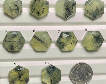 Könnte beinhalten: Zehn sechseckige Prehnit-Edelsteine mit schwarzen Einschlüssen werden gezeigt. Die Steine variieren in der Farbe von blassgelb bis hellgrün. Eine Münze dient zum Größenvergleich. Die Edelsteine sind auf einer weißen Oberfläche angeordnet.