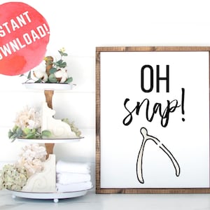 May include: A white wooden sign with a brown frame, featuring the text "OH snap!" in black lettering with a white wishbone illustration.