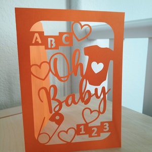 May include: Orange cardstock cut-out card with a window revealing the words "Oh Baby" in white, surrounded by hearts, a baby onesie, a pacifier, the letters "ABC", and the numbers "123".