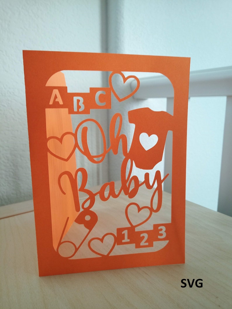 May include: Orange cardstock cut-out card with a window revealing the words "Oh Baby" in white, surrounded by hearts, a baby onesie, a pacifier, the letters "ABC", and the numbers "123".