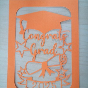 May include: Orange cardstock cut-out design for a graduation card. The design features a graduation cap, diploma, stars, and the text "Congrats Grad 2025".