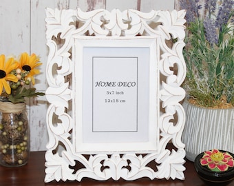 May include: White wooden picture frame with intricate carvings. The frame is 5x7 inches or 13x18 cm. The frame is on a wooden surface.