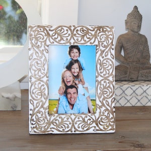 Puede incluir: Marco de fotos de madera blanca con intrincados detalles tallados. El marco contiene una foto de una familia de cuatro personas sonriendo en un campo.