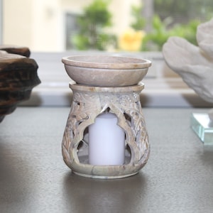 May include: A carved stone oil burner with a white candle inside. The burner has a decorative design of leaves and flowers. The top of the burner is a shallow bowl.
