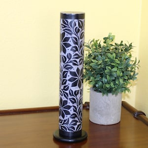 May include: Black and white floral patterned incense burner made of stone. The burner is cylindrical and has a round base. It is sitting on a wooden surface.