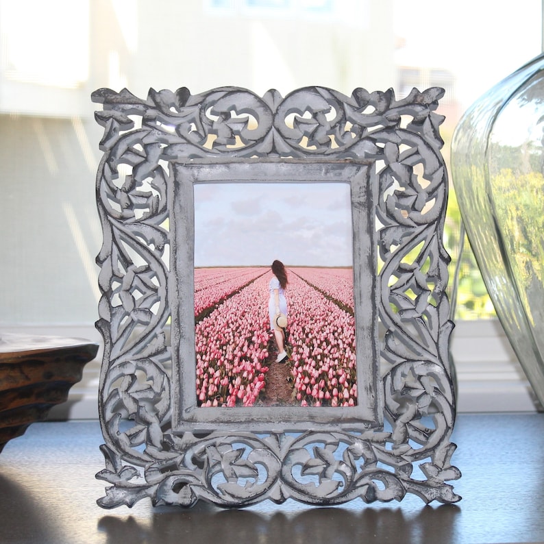 May include: A gray wooden picture frame with an ornate design. The frame holds a photo of a person walking through a field of pink flowers.