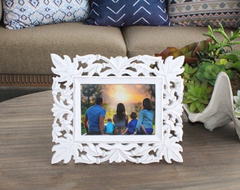 May include: White carved wooden picture frame with a family photo inside. The photo shows a family of four sitting on a bench with a sunset in the background.