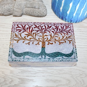 Puede incluir: Una caja rectangular de piedra con un diseño de árbol tallado. El árbol está en tonos de rojo, naranja y verde. La caja está hecha de esteatita y tiene un borde decorativo.