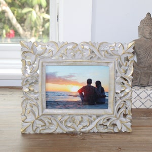 Puede incluir: Marco de fotos de madera blanca con tallas intrincadas y detalles dorados. El marco contiene una foto de una pareja sentada en una playa al atardecer.