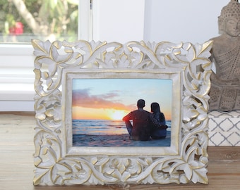 May include: White wooden picture frame with intricate carvings and gold accents. The frame holds a photo of a couple sitting on a beach at sunset.