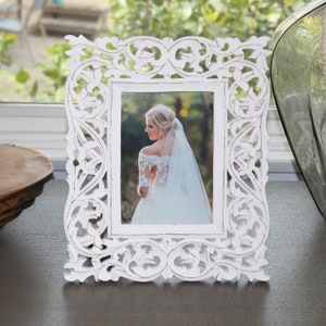 May include: White wooden picture frame with intricate carvings. The frame is holding a photo of a bride in a white wedding dress.