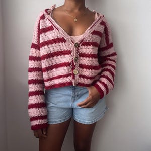 May include: A long-sleeved, cropped crochet cardigan featuring horizontal stripes in alternating shades of light pink and deep red. The garment has a V-neckline, a front button closure with three large, round, light-colored buttons, and long sleeves.