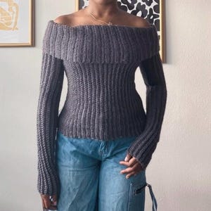 Könnte beinhalten: Ein grauer Off-Shoulder-Pullover mit langen Ärmeln. Der Pullover hat eine gerippte Strickstruktur und einen umgeschlagenen Ausschnitt. Die Ärmel sind eng anliegend und der Körper ist figurbetont. Der Pullover wird mit einer blauen Jeans getragen.