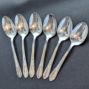 May include: Six silver spoons with a floral design on the handles are arranged on a dark surface. The spoons have a reflective, polished finish, and the bowls of the spoons are oval-shaped.